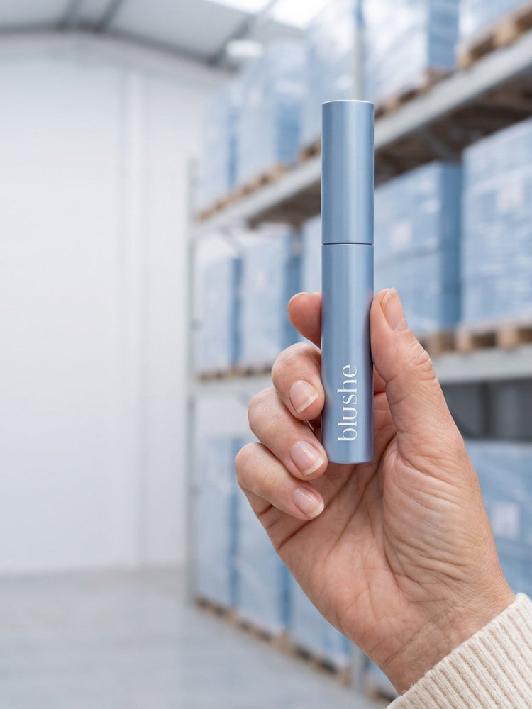 A hand holding a Blushe tube in a warehouse aisle stacked with light blue Blushe boxes